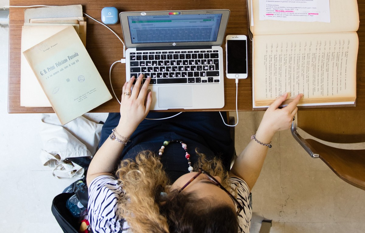 Una studentessa cerca informazioni in un volume antico davanti a un laptop.